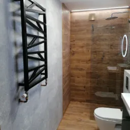 Nowoczesna łazienka z szarymi płytkami na ścianie, drewnopodobnymi płytkami w strefie prysznica, czarnym grzejnikiem drabinkowym i podświetlanym lustrem.