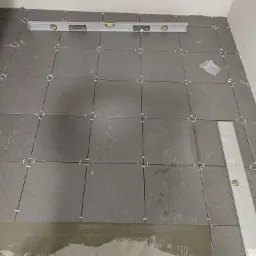 Szare płytki podłogowe ułożone z użyciem krzyżyków dystansowych, poziomica spoczywa na płytkach, widoczny fragment zaprawy klejowej na podłożu.