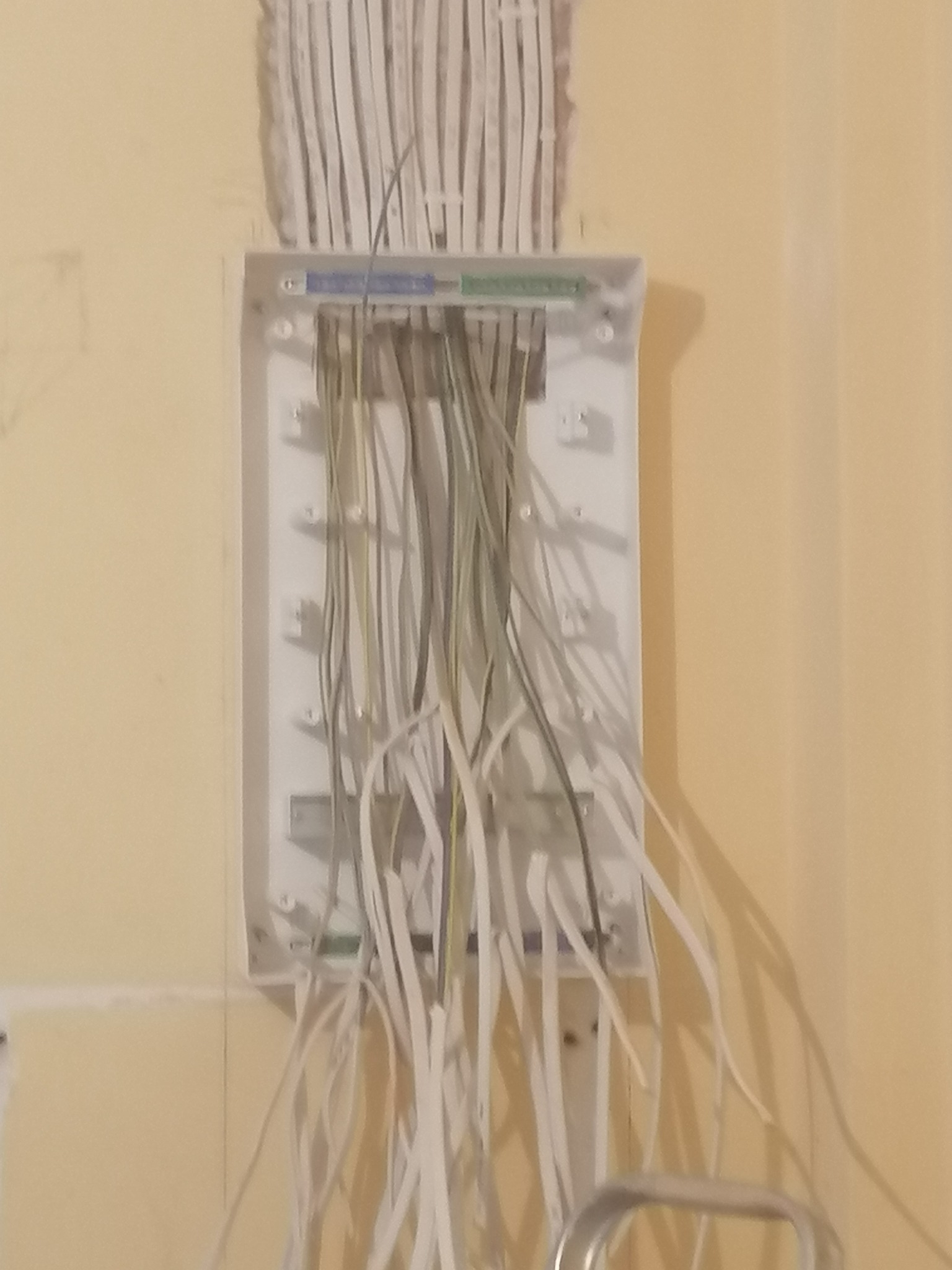 Otwarta puszka elektryczna podtynkowa z wystającymi przewodami w kolorach białym i szarym, osadzona w ścianie o żółtym odcieniu, widoczny fragment drabiny.