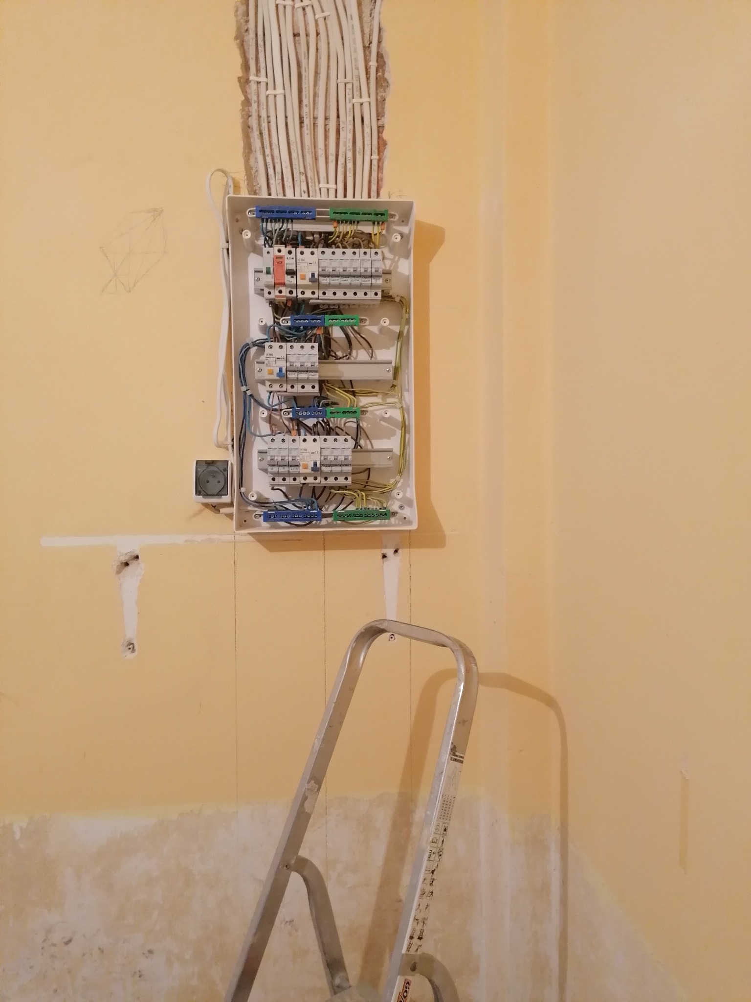 Otwarta skrzynka rozdzielcza z przewodami na pomarańczowej ścianie w trakcie prac, widoczna drabina aluminiowa i ślady po wcześniejszych instalacjach.