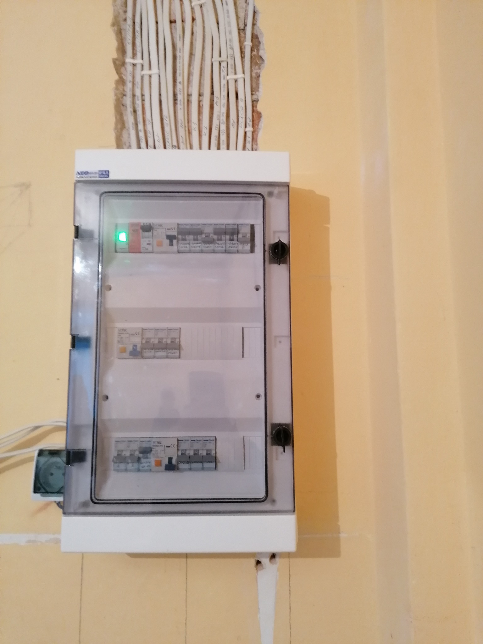 Rozdzielnia elektryczna z widocznymi bezpiecznikami, zamontowana na żółtej ścianie; powyżej wystają kable w peszlach ze ściany, z zieloną diodą sygnalizacyjną.