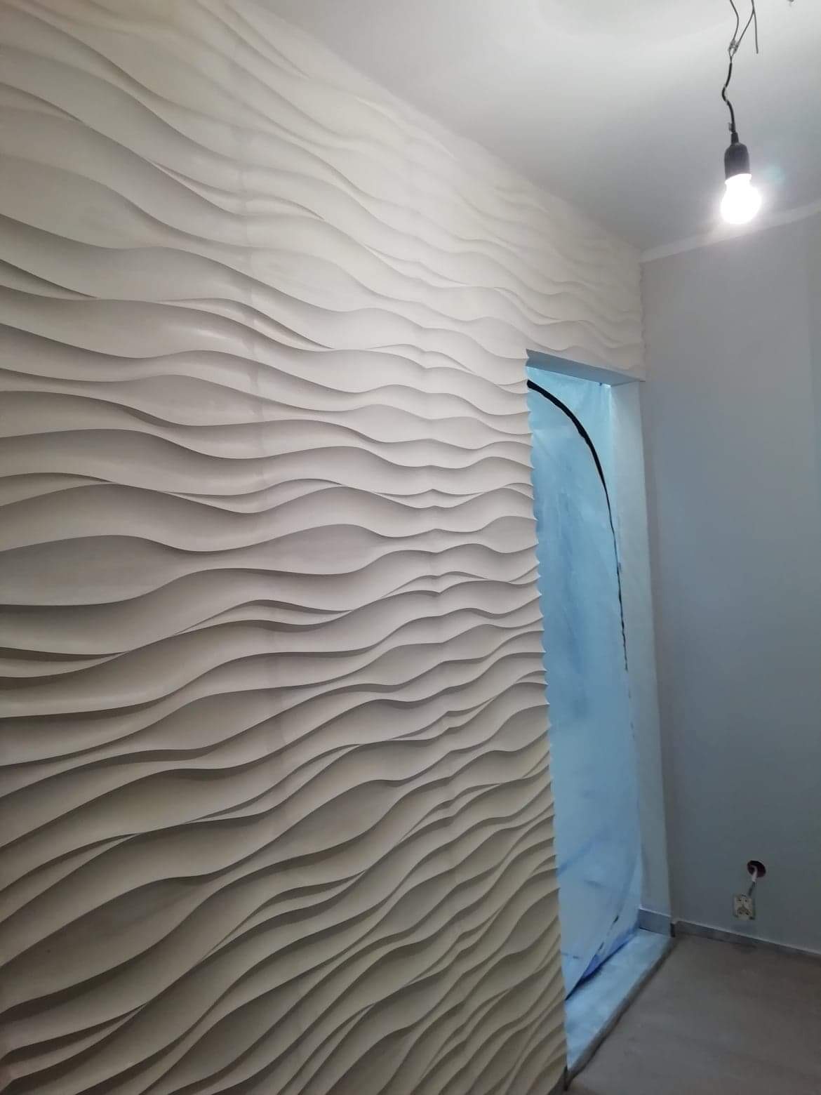 Dekoracyjna ściana z panelami 3D o falistym wzorze w jasnym odcieniu, widok na zabezpieczone folią drzwi w trakcie prac wykończeniowych.