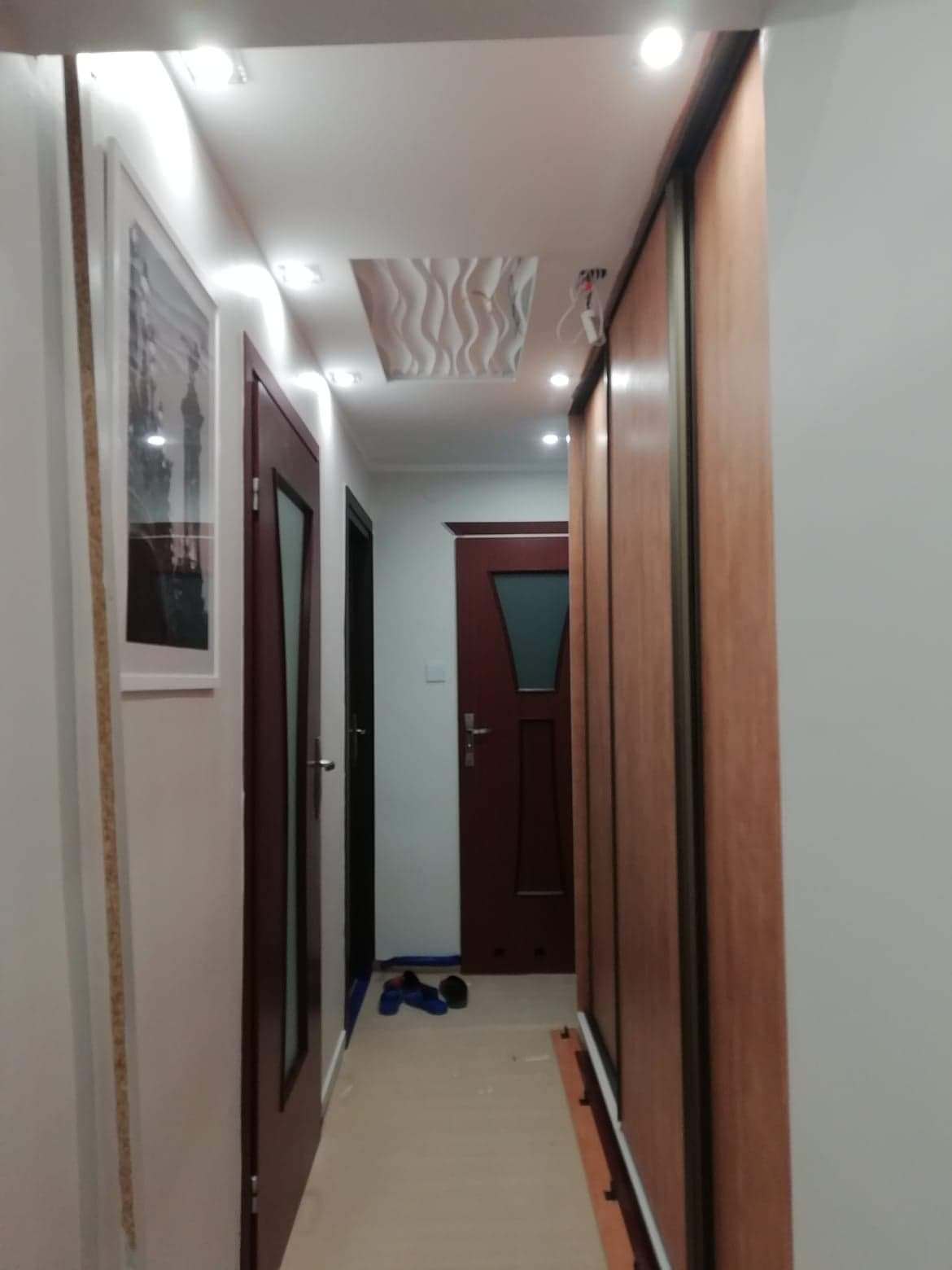 Korytarz po remoncie z przesuwną szafą w zabudowie, dwiema parami drzwi wewnętrznych w kolorze ciemnego drewna, dekoracyjnym panelem sufitowym i obrazem na ścianie.