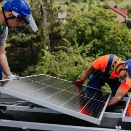 Dwóch monterów w niebieskich kaskach i uprzężach zabezpieczających instaluje panele słoneczne na dachu w Mysłowicach, widok z bliska na pracę w trakcie montażu.
