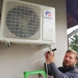 Mężczyzna na zielonej drabince podłącza przewody do białej jednostki klimatyzacji marki Gree zamontowanej na szarej ścianie budynku. Widoczny fragment dachu i drzewa w tle.