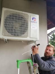 Mężczyzna na zielonej drabince podłącza przewody do białej jednostki klimatyzacji marki Gree zamontowanej na szarej ścianie budynku. Widoczny fragment dachu i drzewa w tle.