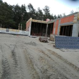 Darex firma budowlana Dariusz Jóźwiak - Budowa domu w stanie surowym otwartym, widoczne ściany z czerwonej cegły, betonowe słupy i przygotowane szalunki, teren budowy z piaskiem i śladami opon, w tle las iglasty.