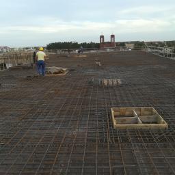 Darex firma budowlana Dariusz Jóźwiak - Siatka zbrojeniowa na płycie fundamentowej budynku w trakcie budowy, widoczni pracownicy w oddali i zabudowa miejska w tle.