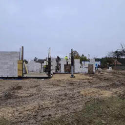 Wznoszenie ścian parterowego budynku z betonu komórkowego. Widoczni pracownicy w odblaskowych kamizelkach, zbrojenie pionowe i szalunki kolumn. Teren budowy przygotowany, pochmurne niebo.