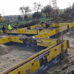 Darex firma budowlana Dariusz Jóźwiak - Budowa fundamentów: szalunki z żółtych desek i zbrojenie pionowe na placu budowy, widoczni pracownicy w oddali.