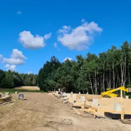 Przygotowanie terenu pod budowę domu: wylane fundamenty, częściowo zasypane piaskiem, widoczny sprzęt budowlany JCB oraz pracownicy na tle lasu i błękitnego nieba z chmurami.