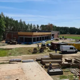 Budowa murowanego budynku z żelbetowymi elementami konstrukcyjnymi, widoczne materiały budowlane, pracownicy w kaskach i pojazdy budowlane na tle zieleni i błękitnego nieba.