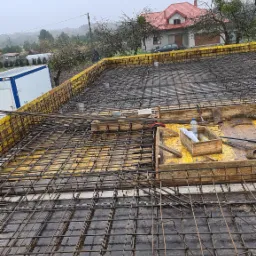 Zbrojenie stropu na żółtym szalunku, widoczne pręty zbrojeniowe ułożone w kratę, drewniane elementy konstrukcyjne, narzędzia w skrzynce, w tle dom z czerwoną dachówką i pochmurne niebo.