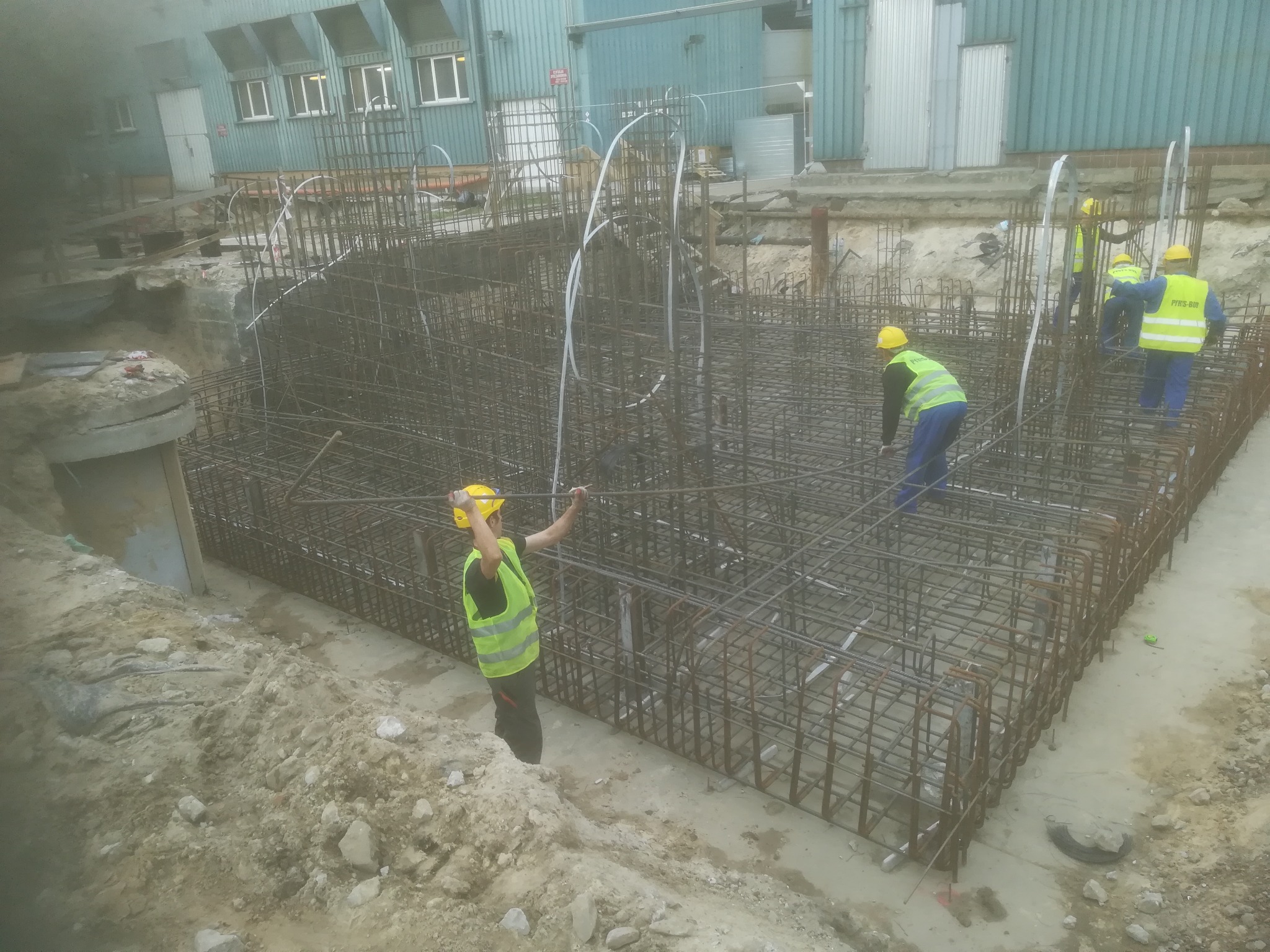 Prace zbrojarskie na budowie: widok na zbrojenie fundamentów z pracownikami w żółtych kamizelkach i kaskach, w tle budynek przemysłowy.