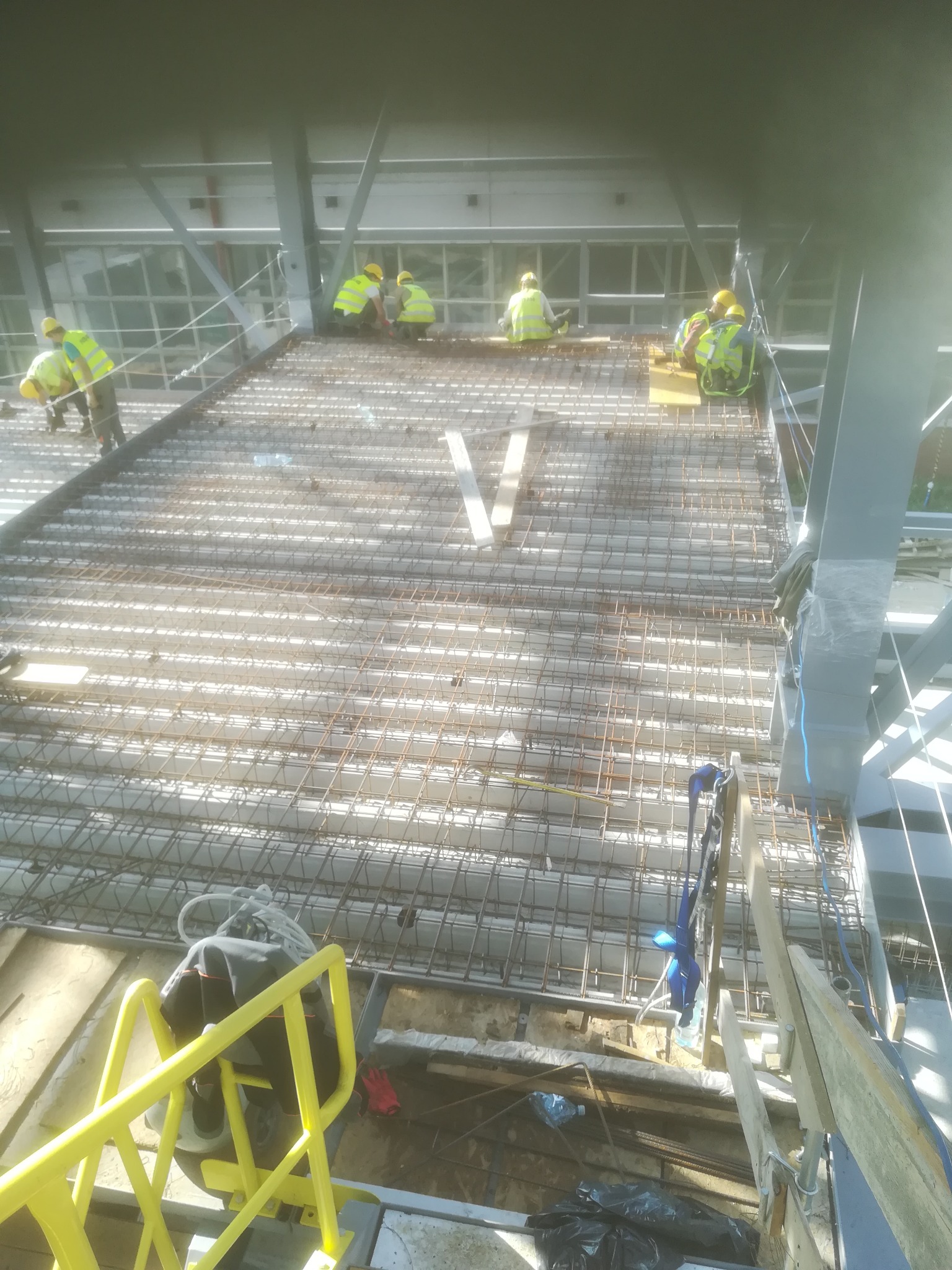 Ekipa budowlana w żółtych kamizelkach i kaskach pracuje na zbrojonej płycie stropowej budynku w trakcie budowy, widok z góry.