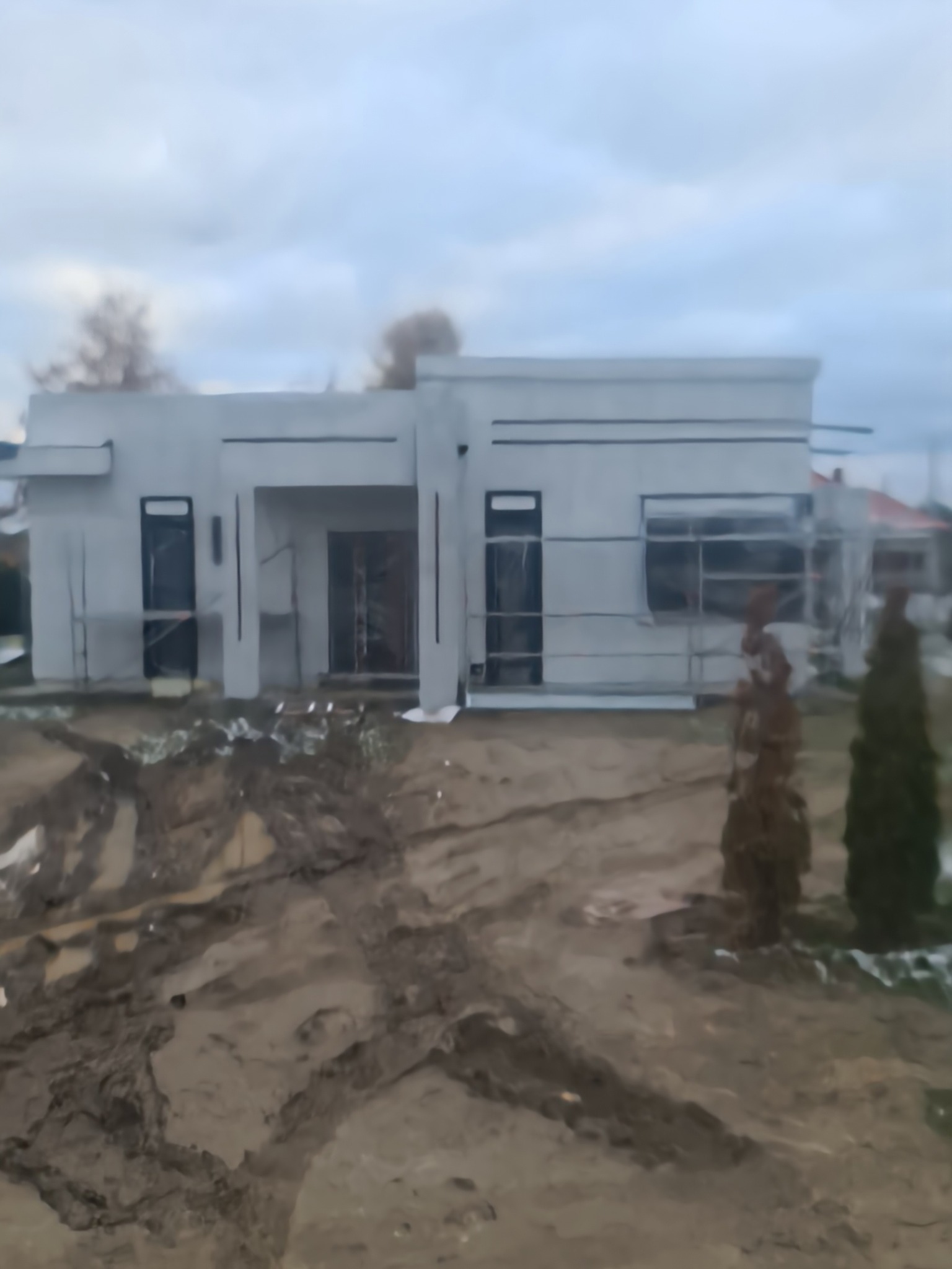 Nowoczesny, parterowy dom w trakcie budowy, z widocznymi rusztowaniami i błotnistym terenem wokół.