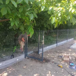 Montaż bramy w ogrodzeniu panelowym na betonowej podmurówce, widoczny pracownik w trakcie instalacji, narzędzia i elementy montażowe rozłożone na ziemi, cień drzew na gruncie.