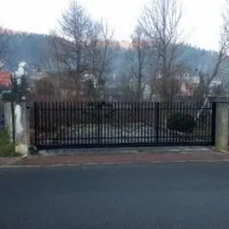 Czarna, przesuwna brama wjazdowa z pionowych prętów, osadzona między dwoma betonowymi słupkami, widok z poziomu ulicy, w tle zabudowa jednorodzinna i zalesione wzgórze.