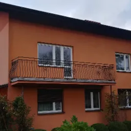 Pomarańczowy dom dwupiętrowy z czarną, metalową balustradą balkonową o dekoracyjnym wzorze. Okna z białymi ramami, częściowo zasłonięte roletami. Widoczne krzewy i drzewa w ogrodzie oraz pochmurne...