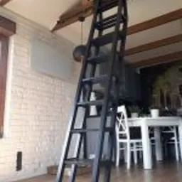 Czarna, metalowa drabina przymocowana do drewnianego sufitu, prowadząca do antresoli w jasnym wnętrzu z białą cegłą na ścianie i drewnianą podłogą.