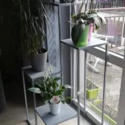 Metalowa konstrukcja regału na kwiaty, w kolorze szarym, z trzema półkami o różnej wysokości, ustawiona przy oknie z widokiem na balkon.