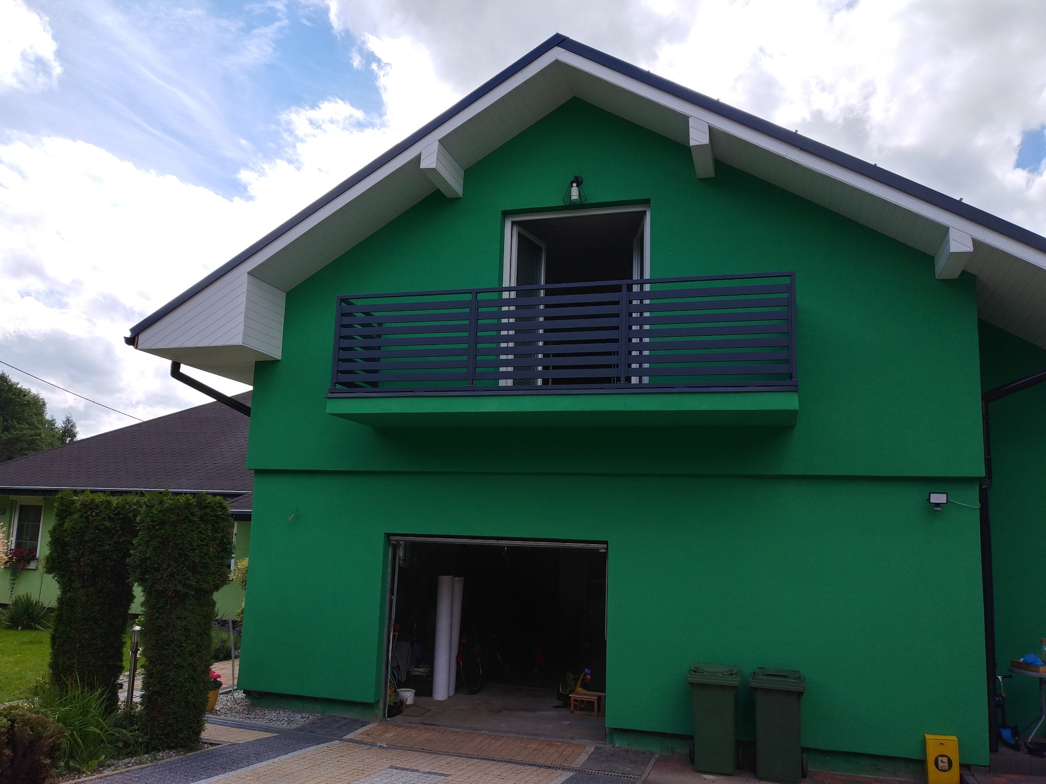 Nowoczesny dom w kolorze zieleni z balkonem z czarną, poziomą balustradą, otwartym garażem i starannie utrzymanymi tujami przy wejściu.