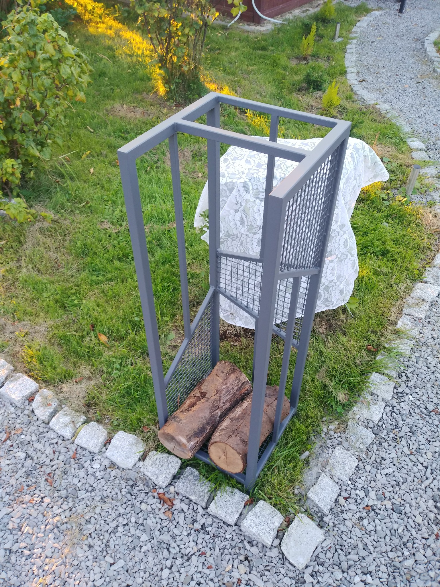 Szary, metalowy stojak na drewno z kratką, ustawiony na trawie obok ścieżki z kamieni.