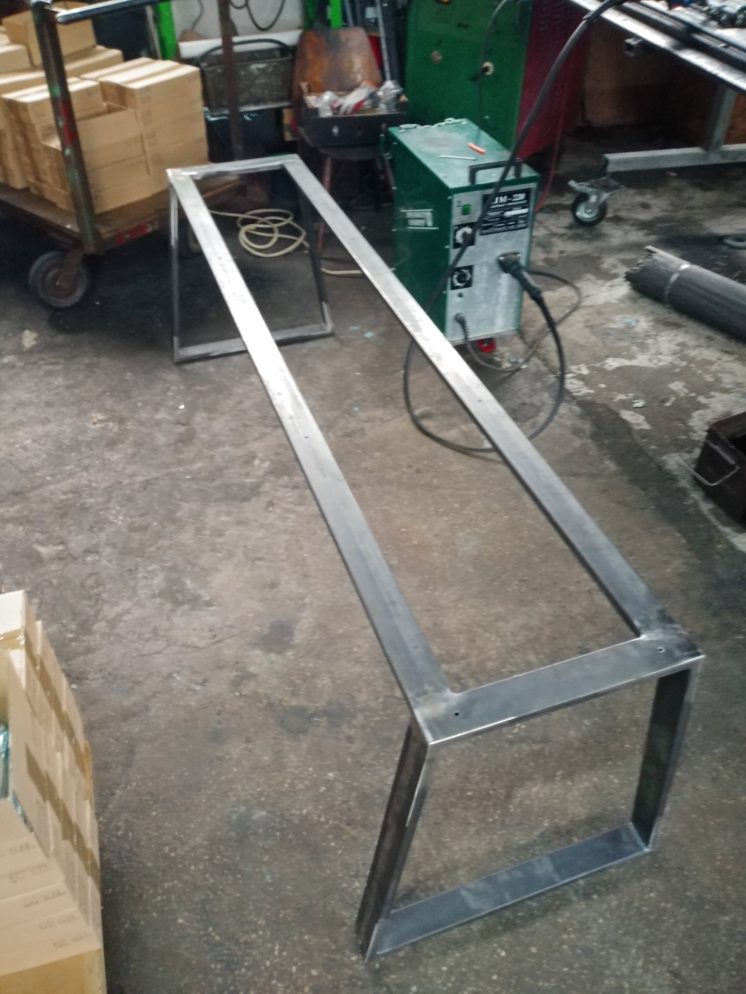 Metalowa rama konstrukcyjna w trakcie spawania, widoczny spawacz, butla z gazem i wózek z kartonami w tle warsztatu.