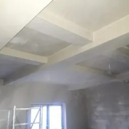 Świeżo nałożony, gładki tynk maszynowy na suficie z widocznymi belkami konstrukcyjnymi, częściowo otynkowane ściany, okno i fragment rusztowania w pomieszczeniu w trakcie remontu.