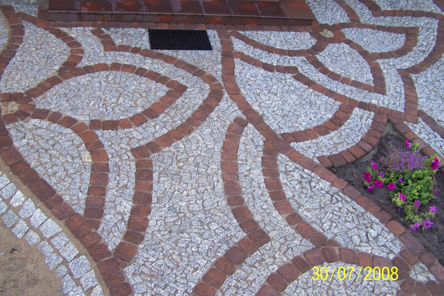 Ułożona nawierzchnia z kostki granitowej i ceglanej, tworząca wzór z zaokrąglonymi liniami, z fragmentem rabaty kwiatowej w prawym dolnym rogu i czarną wycieraczką w tle.