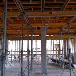 Szare, metalowe podpory stropowe z regulacją wysokości podtrzymujące żółte, drewniane szalunki stropowe z widocznymi oznaczeniami ULMA CC-4 na budowie domu w stanie surowym otwartym.