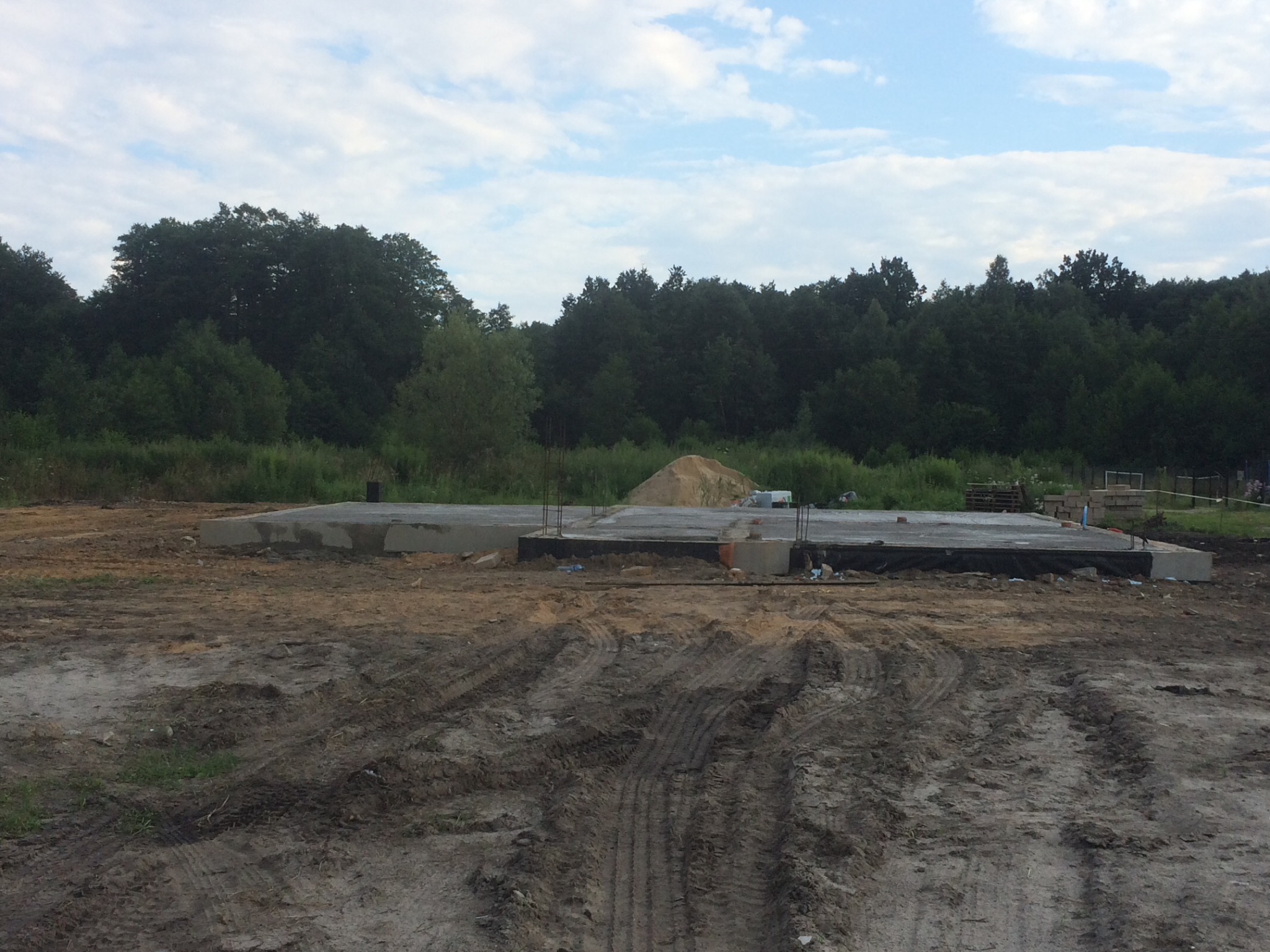 Wylany fundament pod budowę domu jednorodzinnego, widoczne zbrojenia i izolacja, w tle hałda piasku i las.