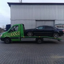 Zielona laweta z czarnym samochodem osobowym marki Audi na platformie, zaparkowana na kostce brukowej przed szarą halą z logo firmy Trilogiq.