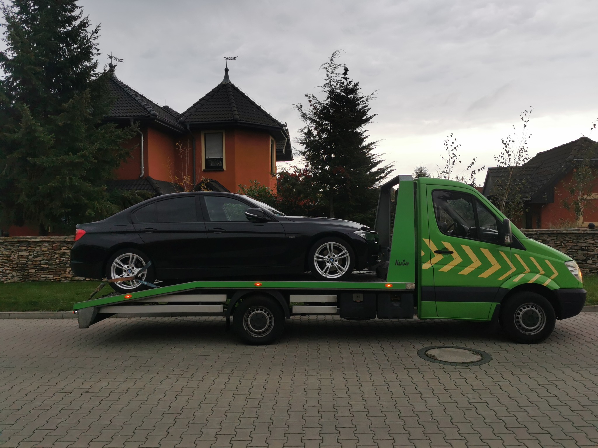 Czarny samochód osobowy marki BMW na zielonej lawecie z żółtymi strzałkami, na tle brukowanej drogi i domu z wieżyczką.