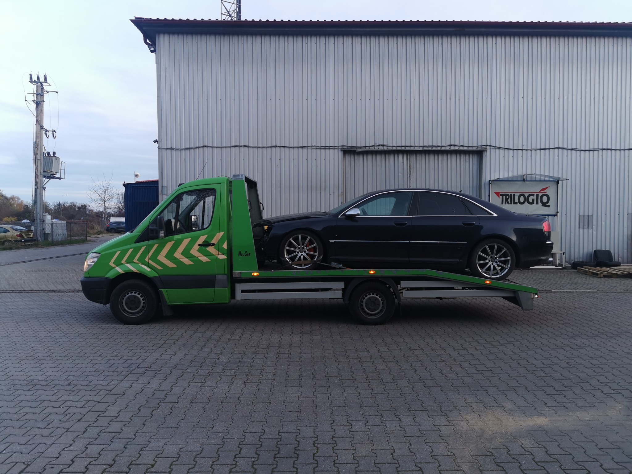 Zielona laweta z czarnym samochodem osobowym marki Audi na platformie, zaparkowana na kostce brukowej przed szarą halą z logo firmy Trilogiq.