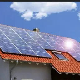 Panele słoneczne na dachu domu jednorodzinnego z czerwoną dachówką, błękitne niebo w tle.