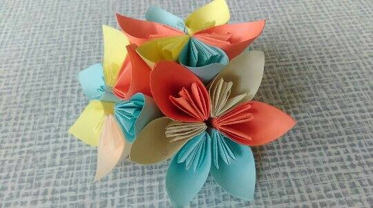 Kompozycja z papierowych kwiatów origami w odcieniach niebieskiego, żółtego, pomarańczowego i beżowego, ułożonych na wzorzystym tle.