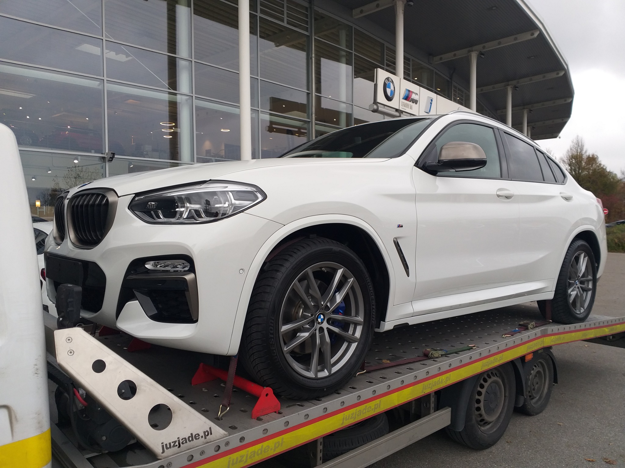 Białe BMW X4 na lawecie transportowej przed salonem samochodowym BMW z widocznym logo firmy.