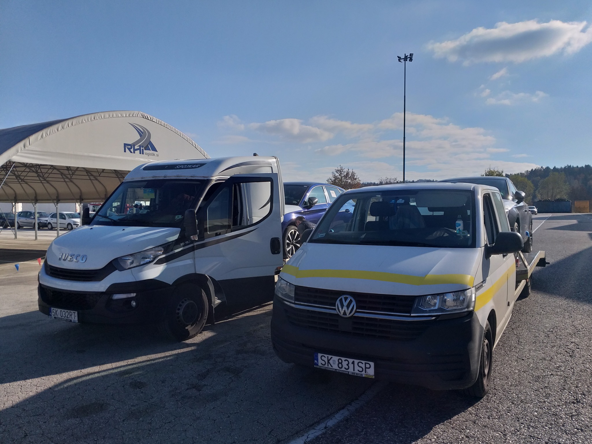 Dwa busy na parkingu, jeden Iveco z czarnymi pasami, drugi Volkswagen z żółtym pasem, w tle hala z logo firmy transportowej, błękitne niebo.