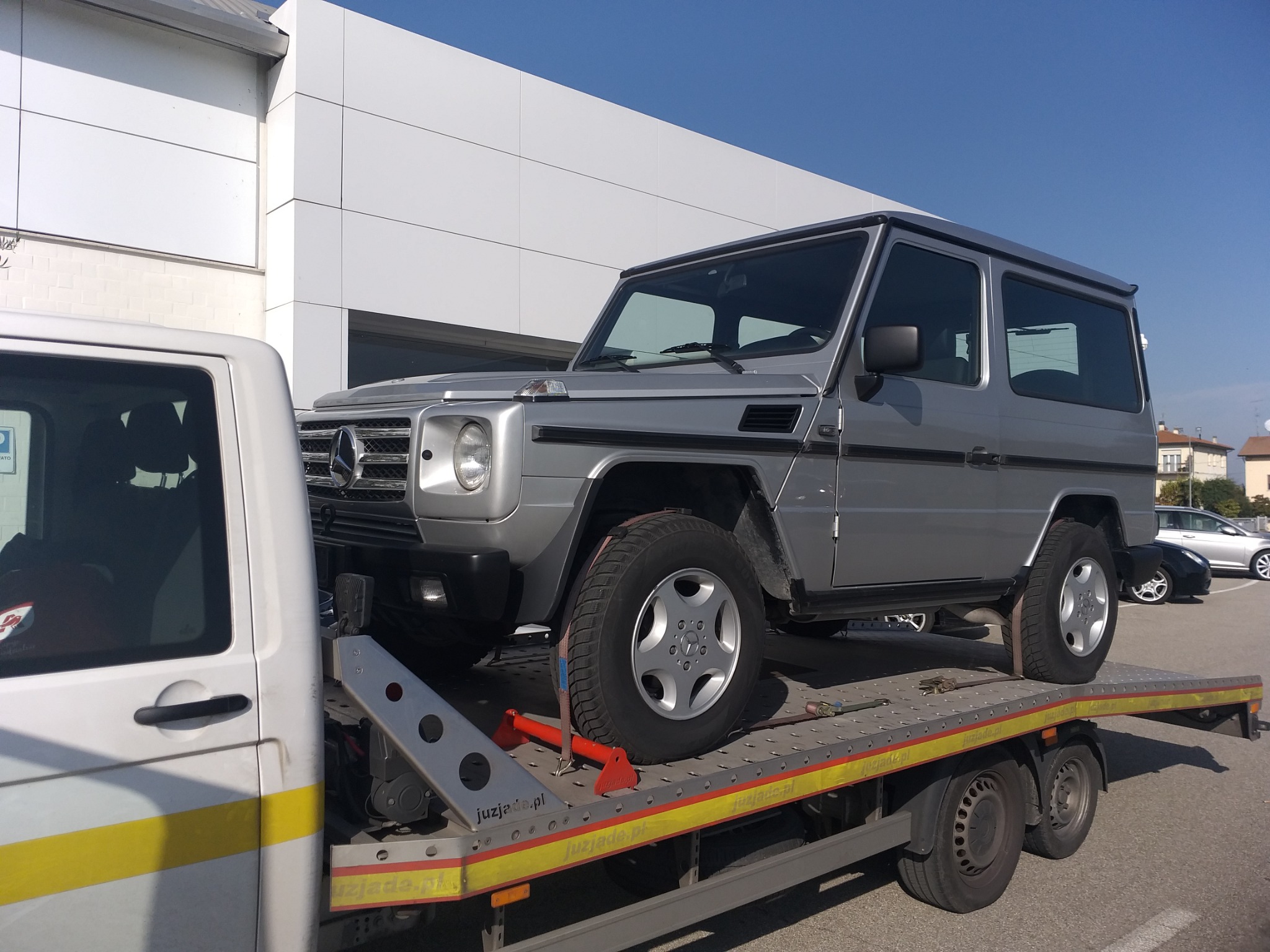 Srebrny Mercedes-Benz G-klasy na platformie lawety transportowej, zabezpieczony pasami i blokadami, na tle białego budynku z panelami.