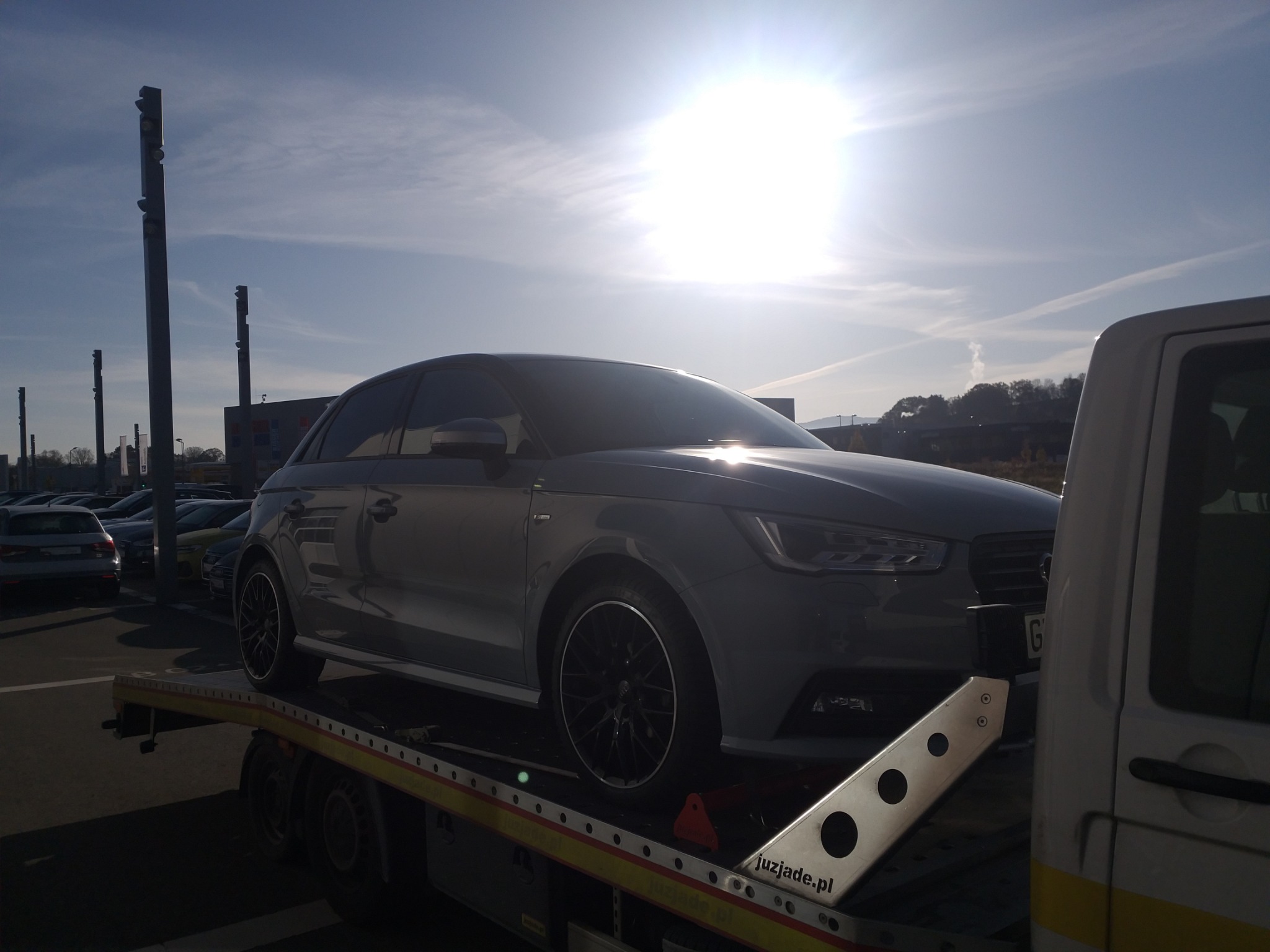 Szary samochód marki Audi A1 na lawecie w słoneczny dzień, widoczne inne auta w tle na parkingu.