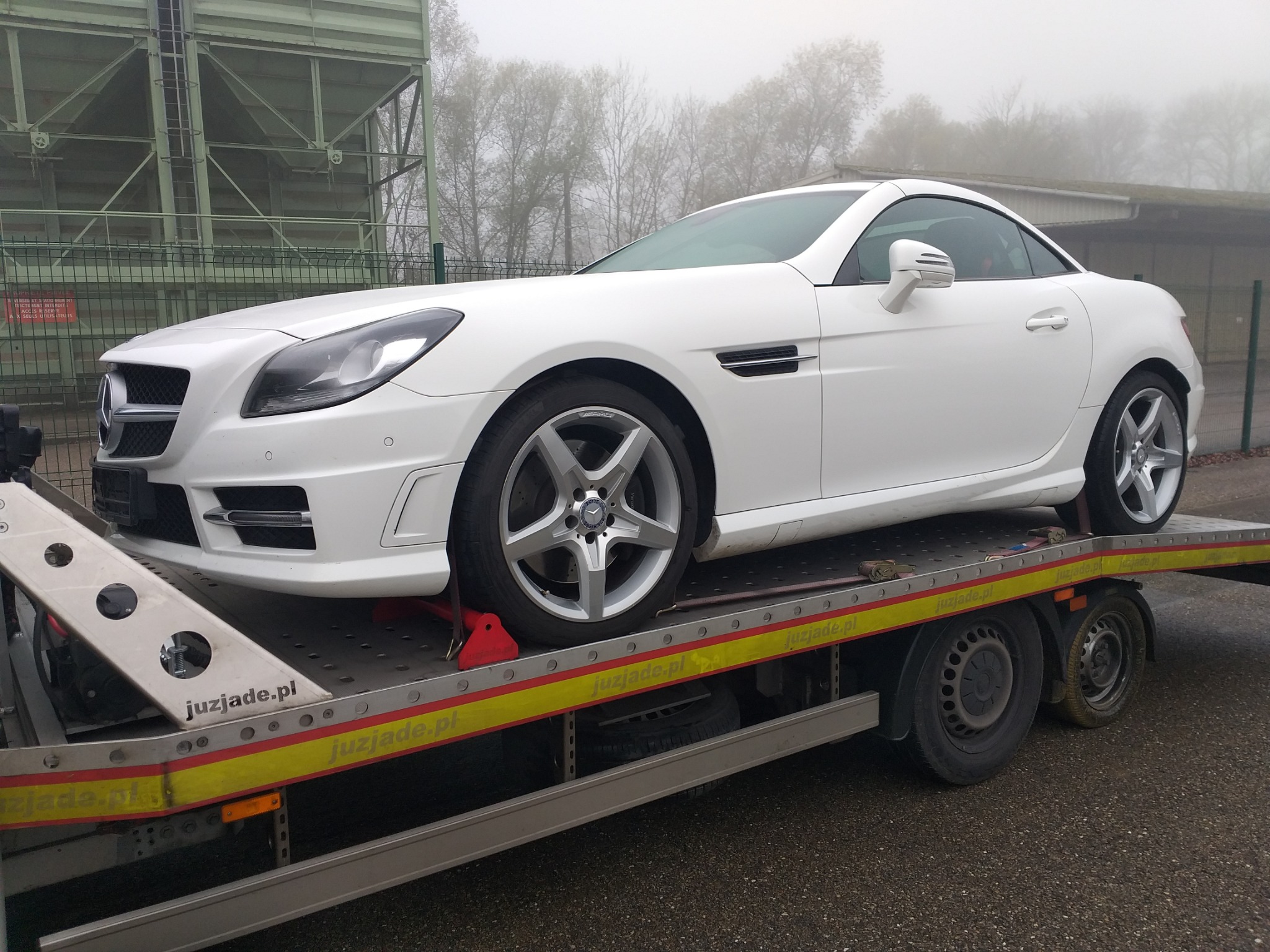 Biały Mercedes SLK na lawecie z czerwonymi zabezpieczeniami kół, w tle zarośla i metalowa konstrukcja w mglisty dzień.