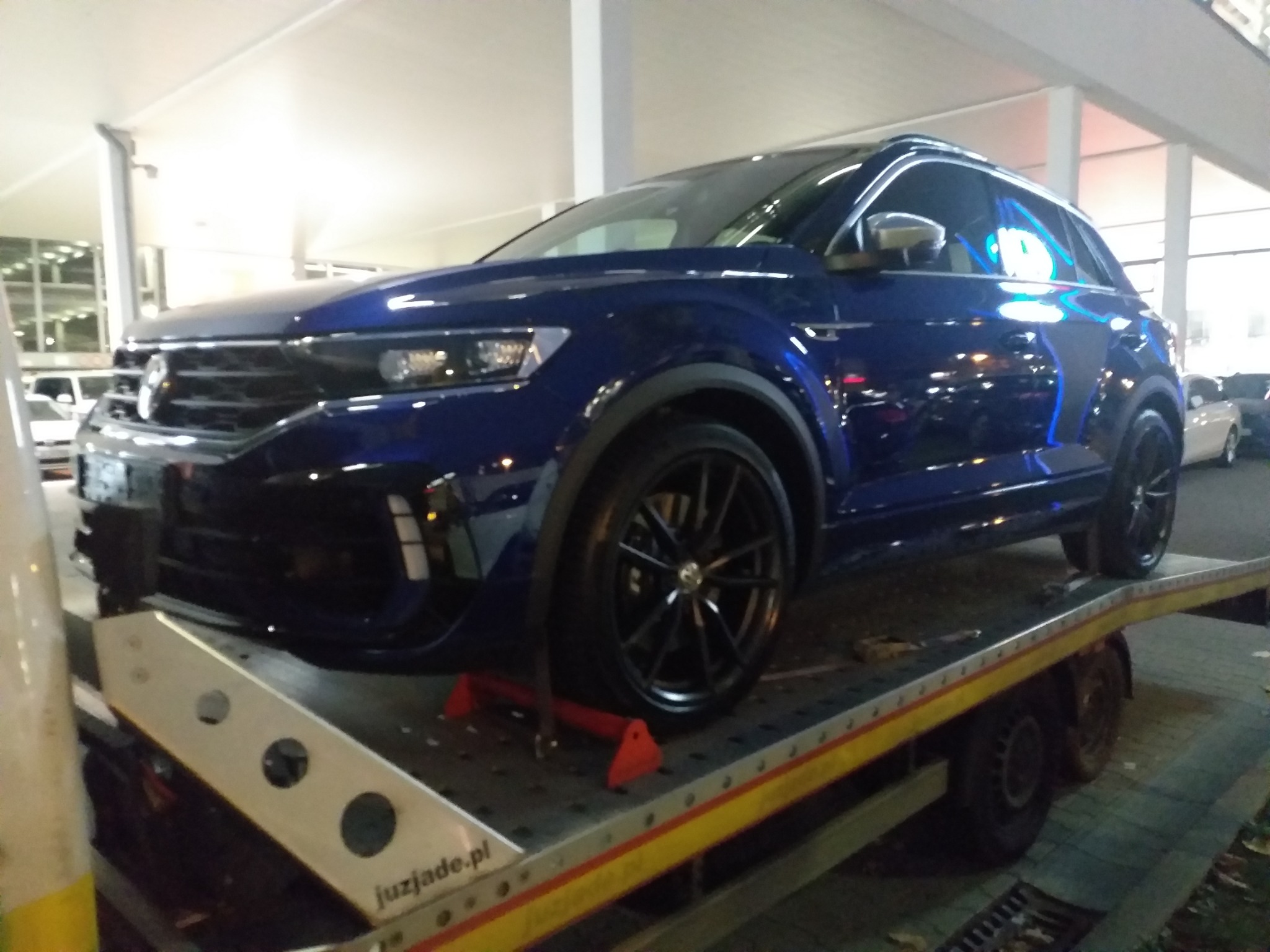 Błyszczący, granatowy Volkswagen T-Roc na platformie lawety z widocznym logo 'juzjade.pl', sfotografowany w słabym oświetleniu miejskim.