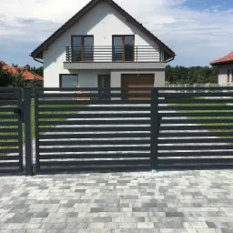 Nowoczesna, pozioma, metalowa brama wjazdowa i furtka w kolorze antracytowym na tle domu z balkonem i kostki brukowej.