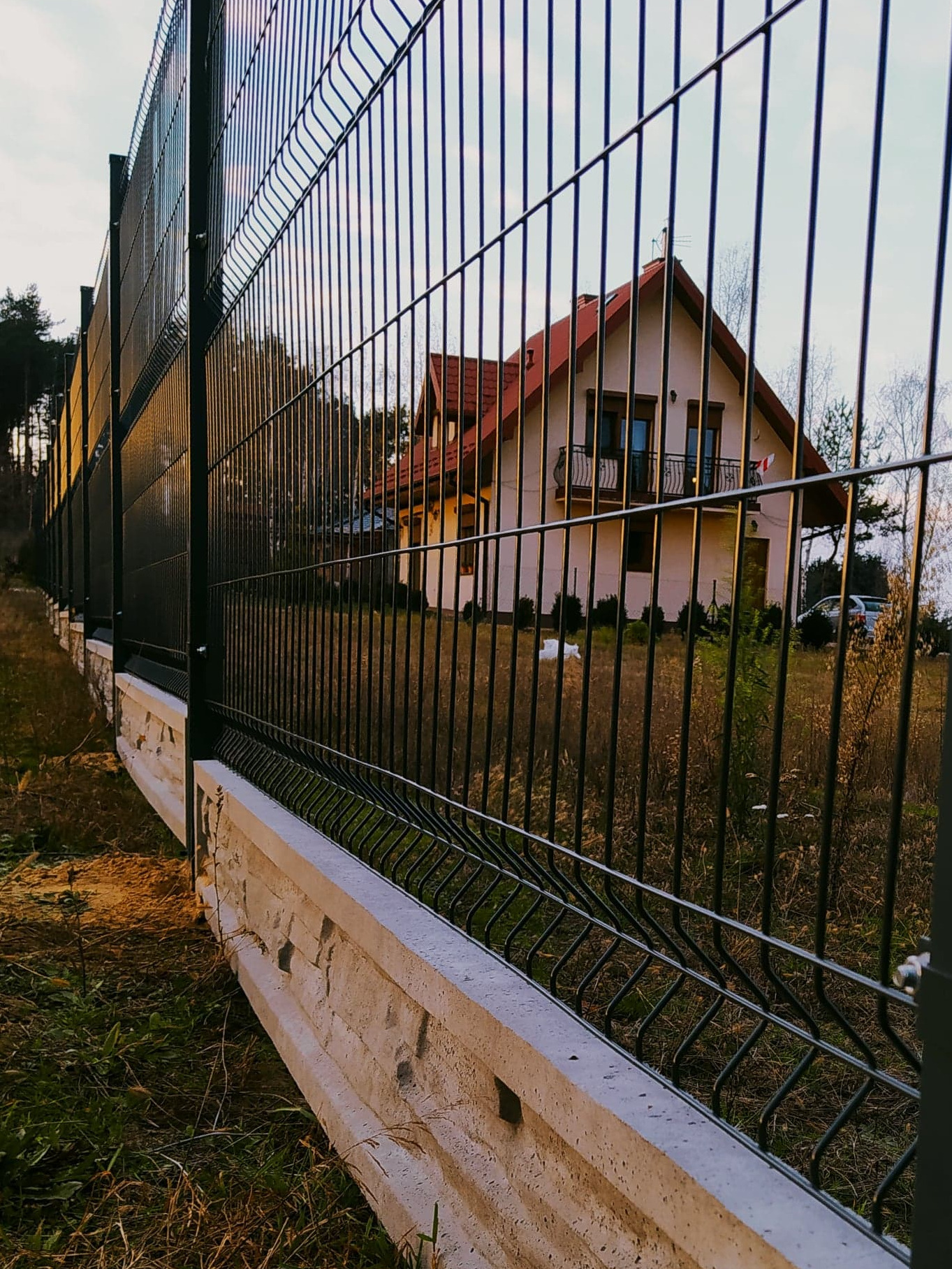 Nowoczesne, czarne ogrodzenie z paneli na betonowej podmurówce, z widokiem na dom z czerwonym dachem i balkonem.