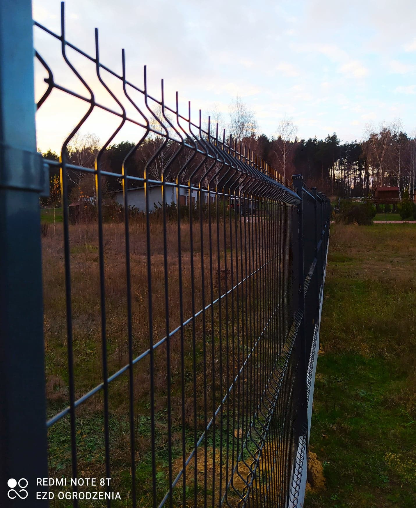 Ciemnoszare ogrodzenie z paneli zgrzewanych na tle łąki i drzew o zmierzchu. Widoczny fragment budynku w oddali.