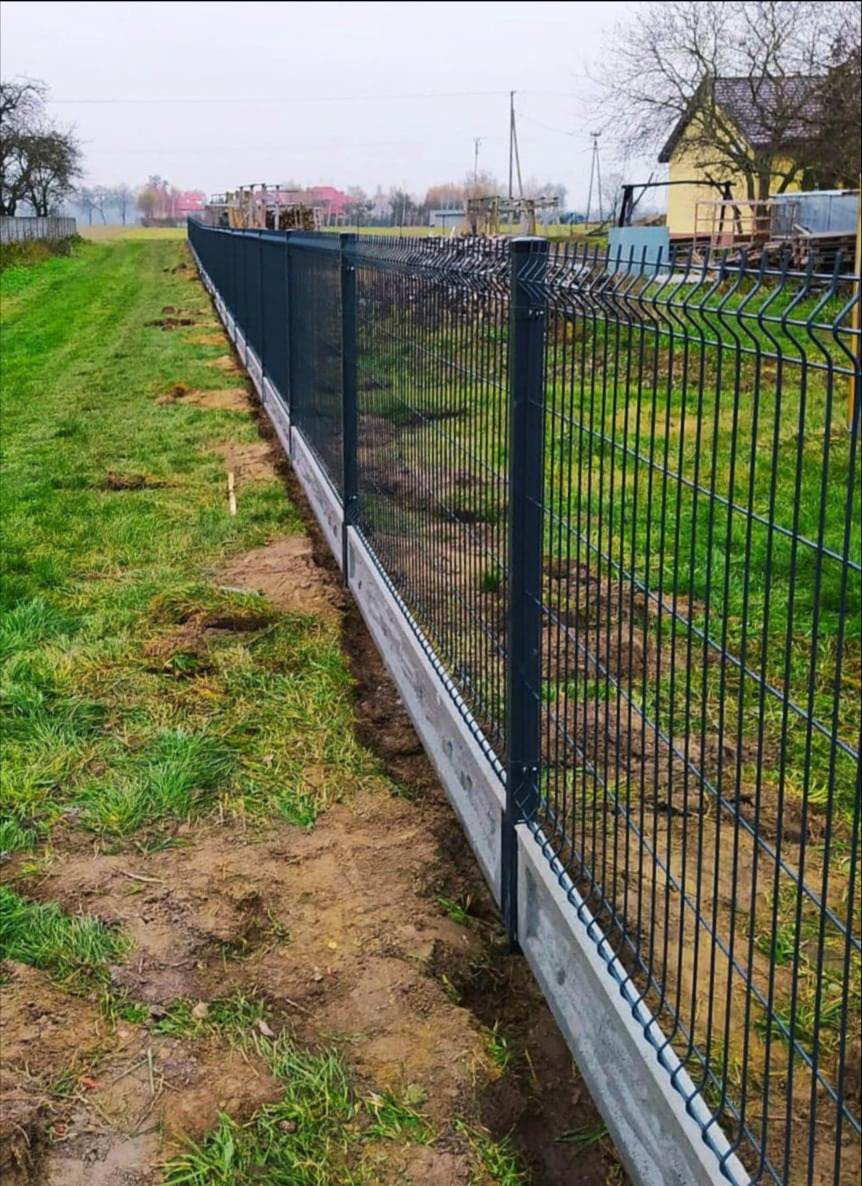 Długie, proste ogrodzenie z paneli zgrzewanych w kolorze grafitowym, osadzone na betonowej podmurówce, oddzielające działkę od łąki.