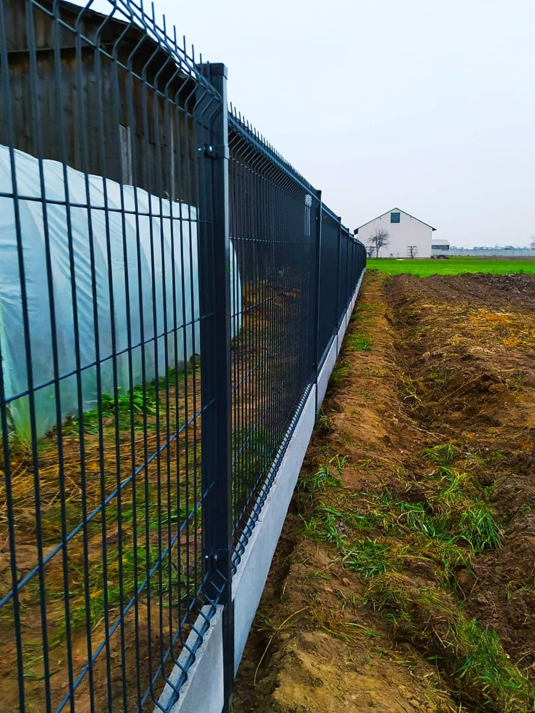 Nowoczesne, grafitowe ogrodzenie z paneli z podmurówką betonową, widok z zewnątrz posesji, w tle budynek gospodarczy i pole.
