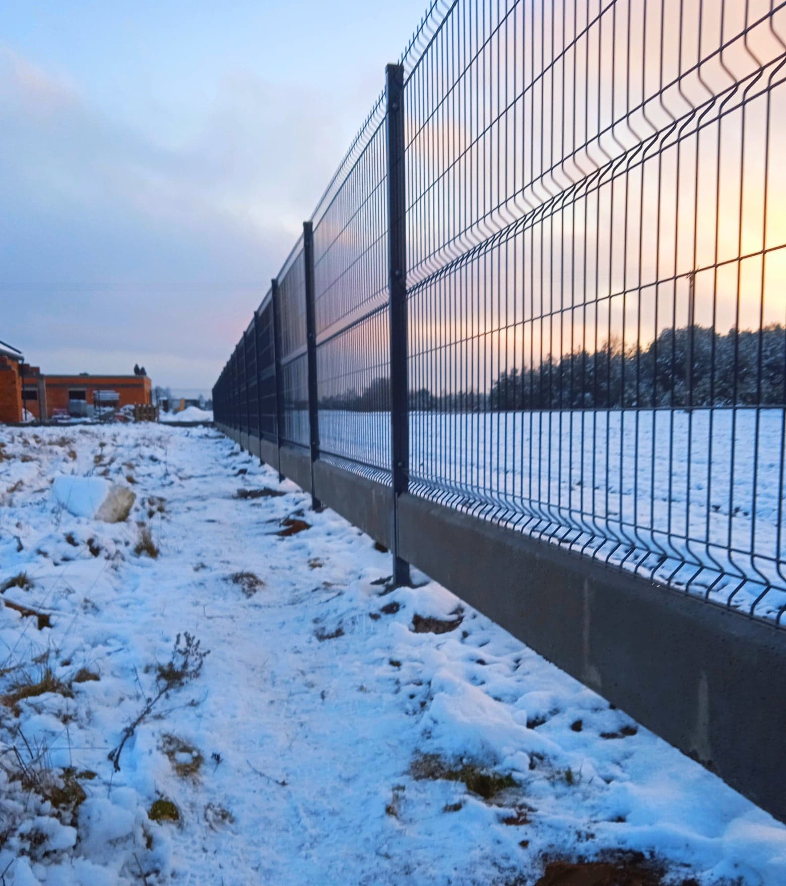 Długie, ciemne ogrodzenie panelowe z podmurówką betonową, widoczne na tle zaśnieżonego terenu i zachodzącego słońca, z budynkiem w trakcie budowy w oddali.