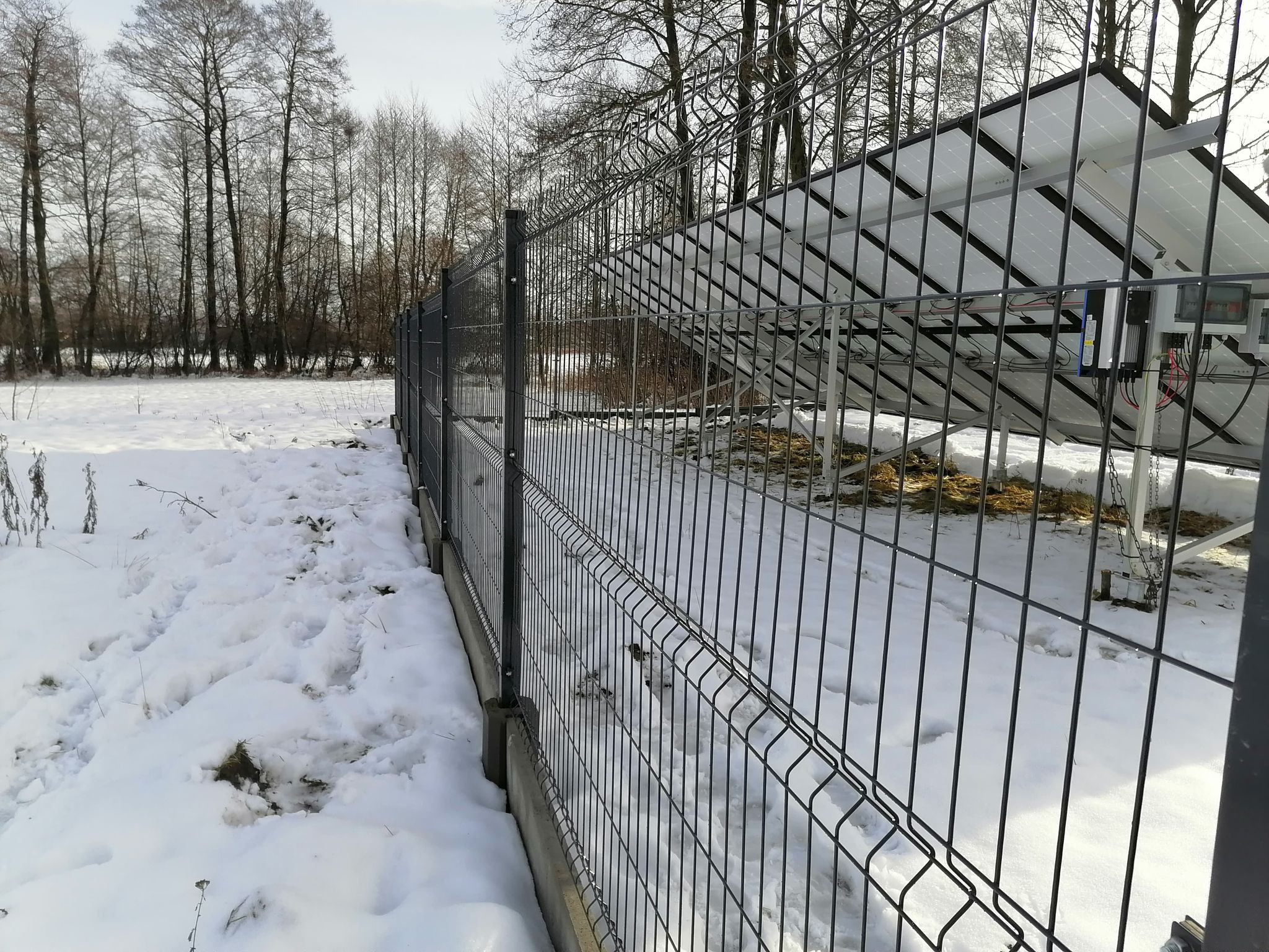 Szare ogrodzenie panelowe z widocznymi panelami fotowoltaicznymi w tle, teren pokryty śniegiem, drzewa bez liści w oddali.
